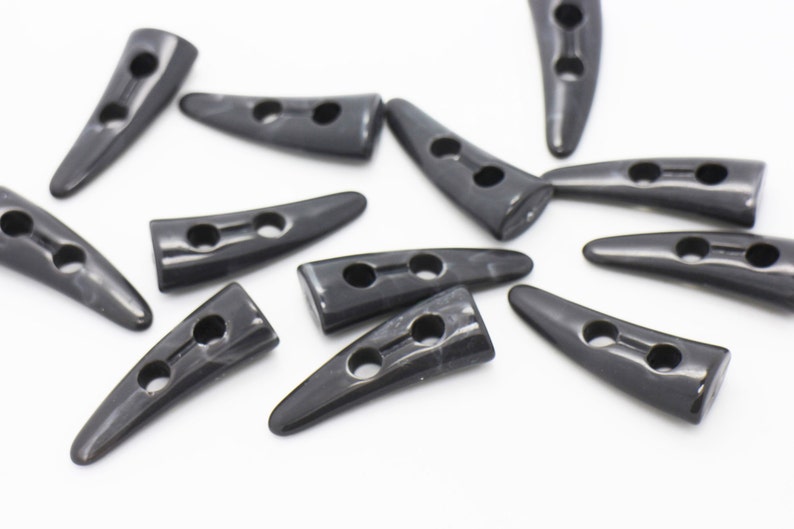 May include: A group of black horn-shaped buttons with two holes each. The buttons are scattered on a white surface.