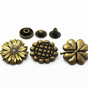 May include: Five antique gold-toned metal buttons with different designs. The buttons feature a daisy, a geometric pattern, a four-leaf clover, and two round buttons with a hole in the centre.