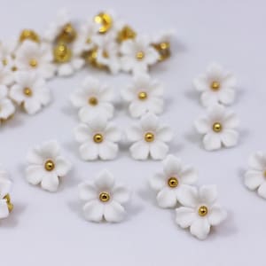 May include: A collection of small white ceramic flowers with gold centers. The flowers are scattered on a white surface.