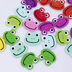 Peut inclure: Un ensemble de boutons en bois de grenouille colorés avec des yeux noirs et des visages souriants. Les boutons sont dans différentes nuances de vert, bleu, jaune, rose, violet et rouge.