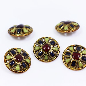 以下が含まれることがあります： 金色の金属フレームとマルチカラーのエナメルデザインが施された装飾ボタン4つ。ボタンは、緑と青の幾何学模様に囲まれた中央の赤い正方形を特徴としています。