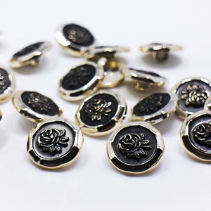 May include: A group of 15 gold and black buttons with a raised rose design. The buttons are scattered on a white surface.