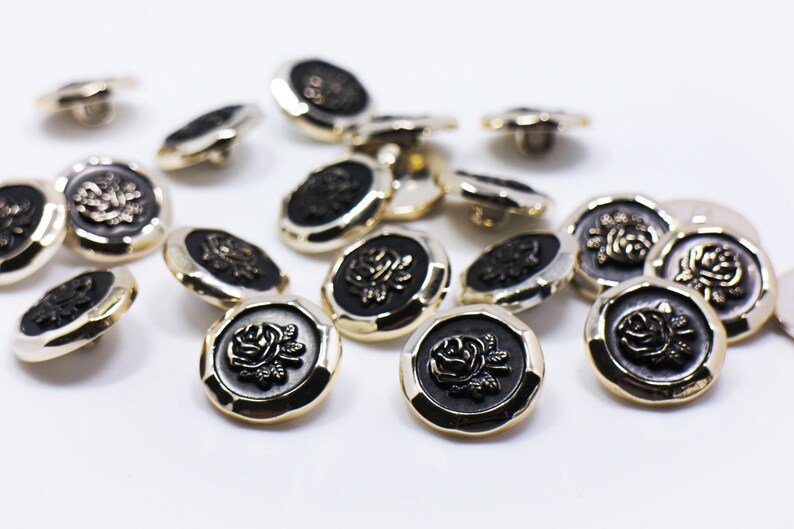 May include: A group of 15 gold and black buttons with a raised rose design. The buttons are scattered on a white surface.