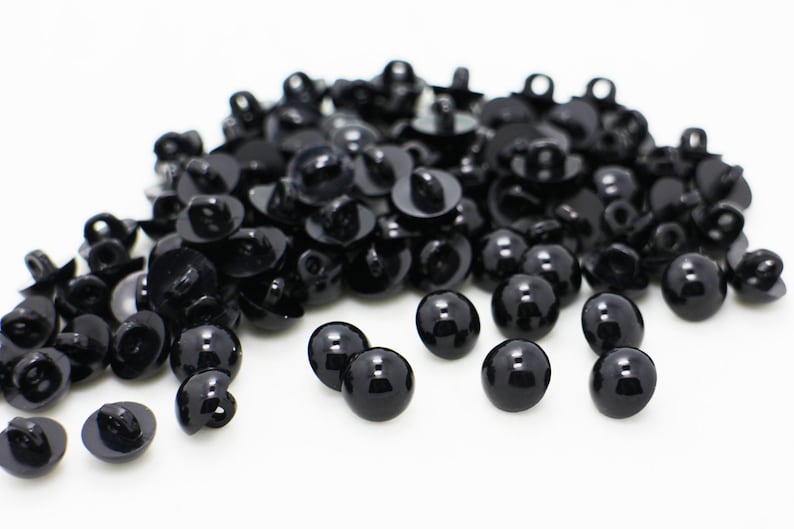 May include: A large pile of black plastic safety eyes for crafts. The eyes are round and have a shiny finish. There are several eyes scattered around the pile.