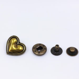 May include: A set of four gold-toned metal heart-shaped snaps. The snaps are used for attaching clothing or other fabric items together.