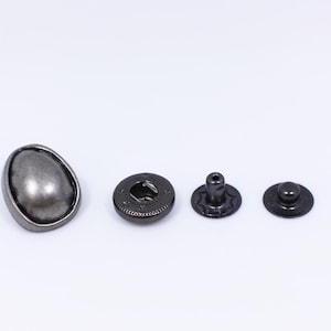 May include: A set of four silver-toned metal snap fasteners. The set includes a dome-shaped button, a round snap with a hole in the center, a post with a star-shaped base, and a round snap with a dome-shaped top.