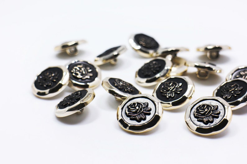 May include: A collection of 14 gold and black buttons with a rose design. The buttons are scattered on a white surface.