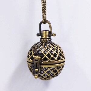 以下が含まれることがあります： 繊細なデザインのゴールドとブラックの金属製のケージペンダントネックレス。ケージは片側が開いており、小さな留め金が付いています。