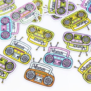 Könnte beinhalten: Eine Sammlung bunter, cartoonartiger Boombox-Patches. Die Patches sind aus Filz und haben eine Vielzahl von Farben, darunter Gelb, Rosa, Blau, Grün und Orange. Die Patches sind perfekt, um jedem Projekt einen Retro-Touch zu verleihen.