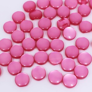 May include: A large group of pink round buttons scattered on a white surface. The buttons are smooth and have a slight shine.