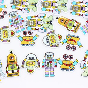 May include: A collection of colorful robot-shaped wooden buttons. The robots feature various designs with different colors, including yellow, blue, green, and purple. They have smiling faces and are scattered on a white surface.