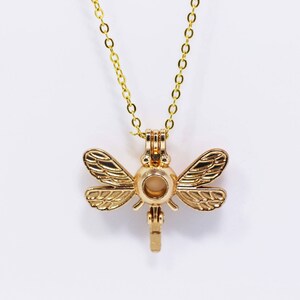Peut inclure: Collier doré avec un pendentif en forme de libellule. La libellule a une ouverture ronde au centre, parfaite pour contenir une petite perle ou un autre charme.