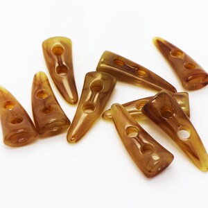 以下が含まれることがあります： 縫い付けるための穴が2つ開いた、角の形をした茶色のプラスチックボタンが8つあります。ボタンは白い表面に散らばっています。