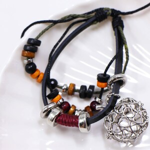 May include: A layered bracelet with black leather cords, wooden beads in black, orange, and brown, and silver-toned metal details. A decorative silver cage charm with heart-shaped cutouts is attached.