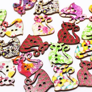 May include: A collection of wooden cat-shaped buttons with various colorful floral and patterned designs. The buttons have two or three holes for sewing and are in shades of green, red, pink, and yellow.