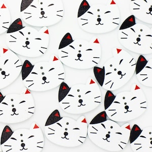 May include: A collection of white wooden buttons with black and red painted cat faces. The buttons are all the same size and shape, and they are arranged in a random pattern.