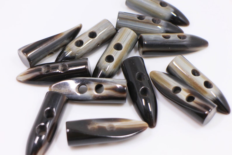May include: A collection of brown and black horn toggle buttons with two holes each. The buttons are in various shapes and sizes, with some being pointed and others being rounded.