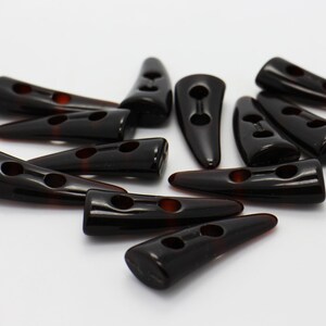 Peut inclure: Un groupe de 12 boutons en plastique noir en forme de corne avec deux trous chacun. Les boutons sont &eacute;parpill&eacute;s sur une surface blanche.