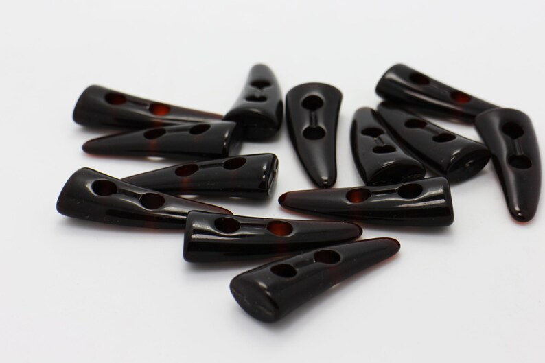 Peut inclure: Un groupe de 12 boutons en plastique noir en forme de corne avec deux trous chacun. Les boutons sont &eacute;parpill&eacute;s sur une surface blanche.