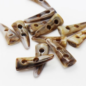 May include: A collection of horn toggle buttons in shades of brown and cream, scattered on a white surface. Each button has two or three holes for sewing. The buttons have a smooth, polished finish and a slightly curved shape.