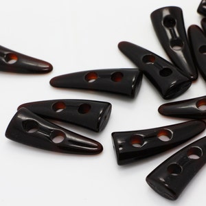 Peut inclure: Un groupe de boutons en plastique brun en forme de corne avec deux trous chacun. Les boutons sont &eacute;parpill&eacute;s sur une surface blanche.