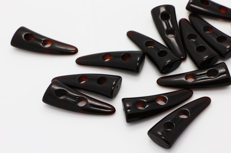 Peut inclure: Un groupe de boutons en plastique brun en forme de corne avec deux trous chacun. Les boutons sont &eacute;parpill&eacute;s sur une surface blanche.