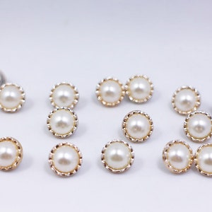 May include: A selection of pearl-effect buttons with gold-coloured scalloped edges. The round buttons have a creamy white colour and a shiny surface. These buttons are likely for sewing or craft projects.