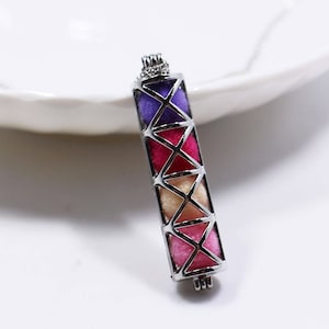May include: A silver pendant with a geometric design. The pendant is rectangular with colourful, triangular sections in shades of purple, pink, and beige. The pendant is on a white surface.