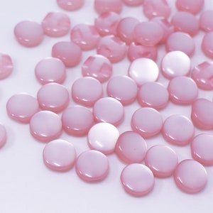 May include: A collection of pink, round, pearl-like buttons. The buttons are scattered on a white surface.