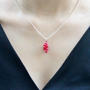 Può includere: Una collana a catena d'argento con un ciondolo di perline rosse. Il ciondolo ha la forma di un grappolo di piccole perline rotonde.