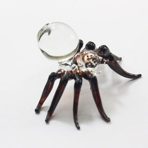 May include: A glass spider figurine with a clear glass orb on its back. The spider has eight legs and a brown body.