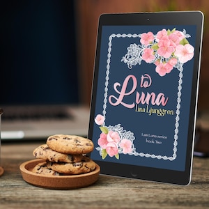 May include: A light blue enamel mug with coffee sits on a wooden surface next to a small plate of chocolate chip cookies. A tablet displays the book cover "to Luna" by Lina Ljunggren, with pink floral accents.
