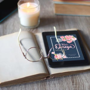 May include: An open book with a pair of round glasses resting on the pages. A digital tablet displaying a floral design with the text "From Omega" is on the right page. A lit candle and a smartphone are in the background.
