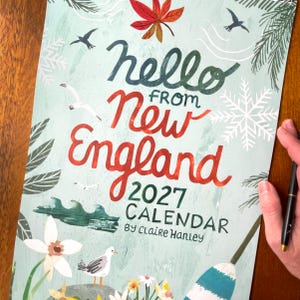 May include: A 2027 calendar titled Hello From New England by Claire Hanley, featuring a light green background with illustrations of a red maple leaf, seagulls, pine branches, snowflakes, waves, and flowers, with a pen resting on the side.