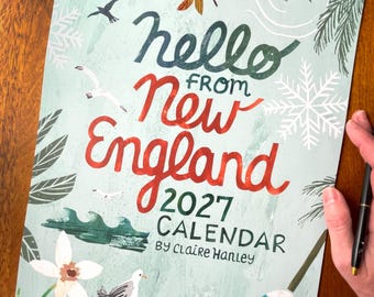 May include: A 2027 calendar titled Hello From New England by Claire Hanley, featuring a light green background with illustrations of a red maple leaf, seagulls, pine branches, snowflakes, waves, and flowers, with a pen resting on the side.
