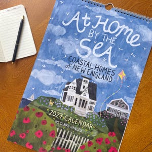 May include: A 2027 wall calendar titled At Home by the Sea Coastal Homes of New England by Claire Hanley. The cover features a blue sky with white clouds, a white house, a kite, a dog, and red flowers. A notebook and pen are placed nearby.