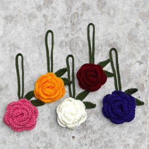 May include: Five crocheted rose ornaments in pink, orange, white, dark red, and purple. Each flower features a dark green crocheted stem with a loop at the top for hanging, and two small dark green crocheted leaves attached to the base of the rose.