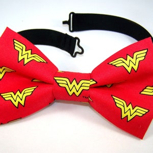 Może przedstawiać: Czerwony krawat w kształcie muszki z powtarzającym się wzorem żółtych logo Wonder Woman.