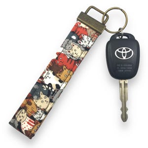 May include: A key chain with a brown fabric strap featuring a repeating pattern of colorful cartoon cats. The strap is attached to a silver key ring with a black car key fob. The key fob has the text "FCC ID: HYD7280M IC: 1091A-T280M 1280M DENSO".