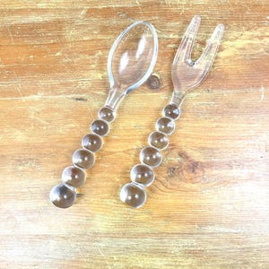 May include: Clear plastic serving spoon and fork set. The handles are made of a series of clear, round beads. The spoon has a rounded bowl, and the fork has three tines. The set is displayed on a wooden surface.
