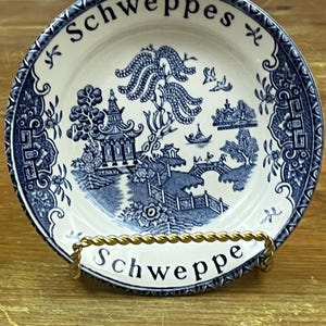 May include: A blue and white ceramic plate with a willow pattern design. The plate has a gold rim and the word "Schweppes" written on it in black lettering. The plate is on a gold stand.