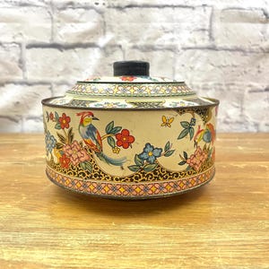 Op de afbeelding: Een vintage, ronde blikken container met een deksel, met een crèmekleurige achtergrond en kleurrijke bloemen- en vogelillustraties. De container heeft een zwarte knop aan de bovenkant en decoratieve rand.