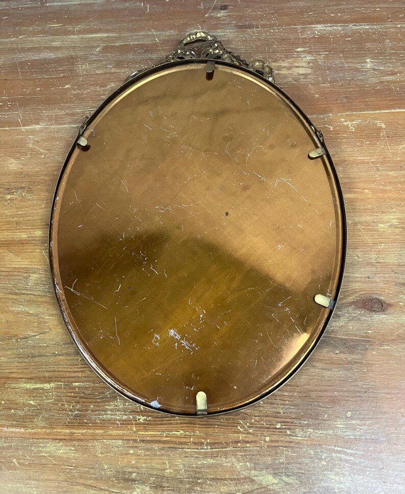 May include: The back of an oval-shaped brass picture frame with a decorative design around the edge. The frame is made of a shiny, gold-colored metal.