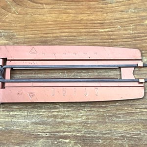 May include: A vintage, copper-colored metal device with the word "DEVILS" embossed on it. The device has a hinged design with two parallel black bars and triangular markings. It rests on a wooden surface.