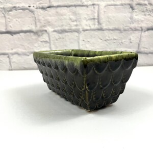 May include: A green glazed ceramic planter with a textured, scale-like design. The planter is rectangular in shape and has a slightly rounded bottom.