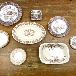 May include: A collection of antique brown and white ceramic dishes, including plates, a serving platter, and a small rectangular dish. The plates feature decorative patterns and scenes. The dishes are arranged on a wooden surface.