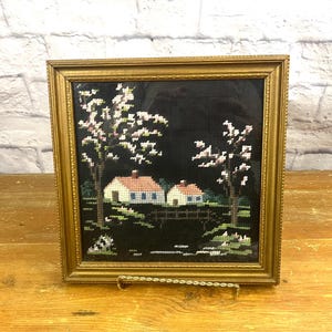 Op de afbeelding: Een ingelijst naaldkunstwerk met twee huizen en bloeiende bomen. De huizen zijn wit met blauwe versieringen en de bomen zijn wit met roze bloemen. De achtergrond is zwart.