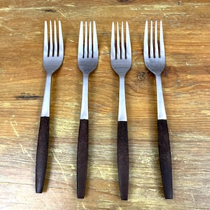 May include: Four stainless steel forks with dark brown wooden handles. The forks are arranged horizontally on a wooden surface. Each fork has four tines and a silver neck connecting the tines to the handle.