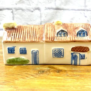 Op de afbeelding: Een keramieken huisje met een crèmekleurige buitenkant en blauw geschilderde ramen en deuren. Het dak is getextureerd met bruine verticale lijnen en bevat twee dakkapellen. Een groen geschilderde struik bevindt zich bij de voordeur.