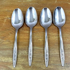 May include: Four silver spoons with floral patterned handles are displayed on a wooden surface. The spoons have a polished, reflective finish, and the spoon bowls are oval-shaped.
